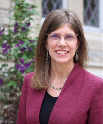 Dr. Mary Stegmaier in a fuchsia blazer standing in front of purple flowers, light brown hair, wearing glasses. 